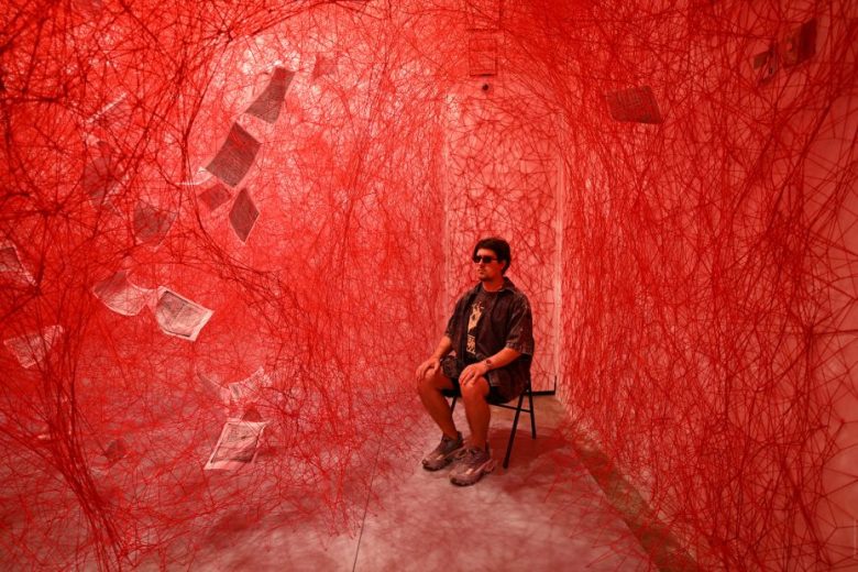 A person sits on a chair in a room filled with dense red string webs and suspended sheets of paper, under red lighting.