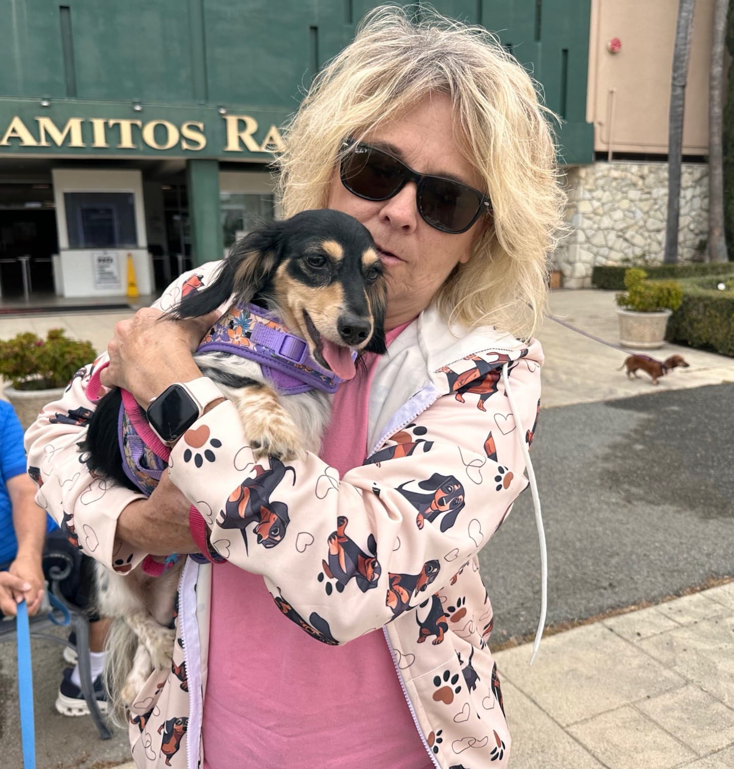 A woman wearing sunglasses cradles a small black and beige dog.