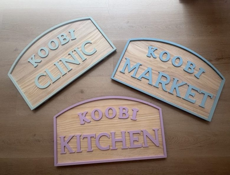 Three wooden signs on a table read "KOOBI CLINIC," "KOOBI MARKET," and "KOOBI KITCHEN," each with colored borders.