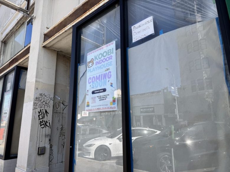 Storefront window with a poster reading "KOOBI INDOOR PLAYHOUSE COMING SOON" and a handwritten sign above saying "Business Coming Soon"; cars and buildings reflected in the glass.