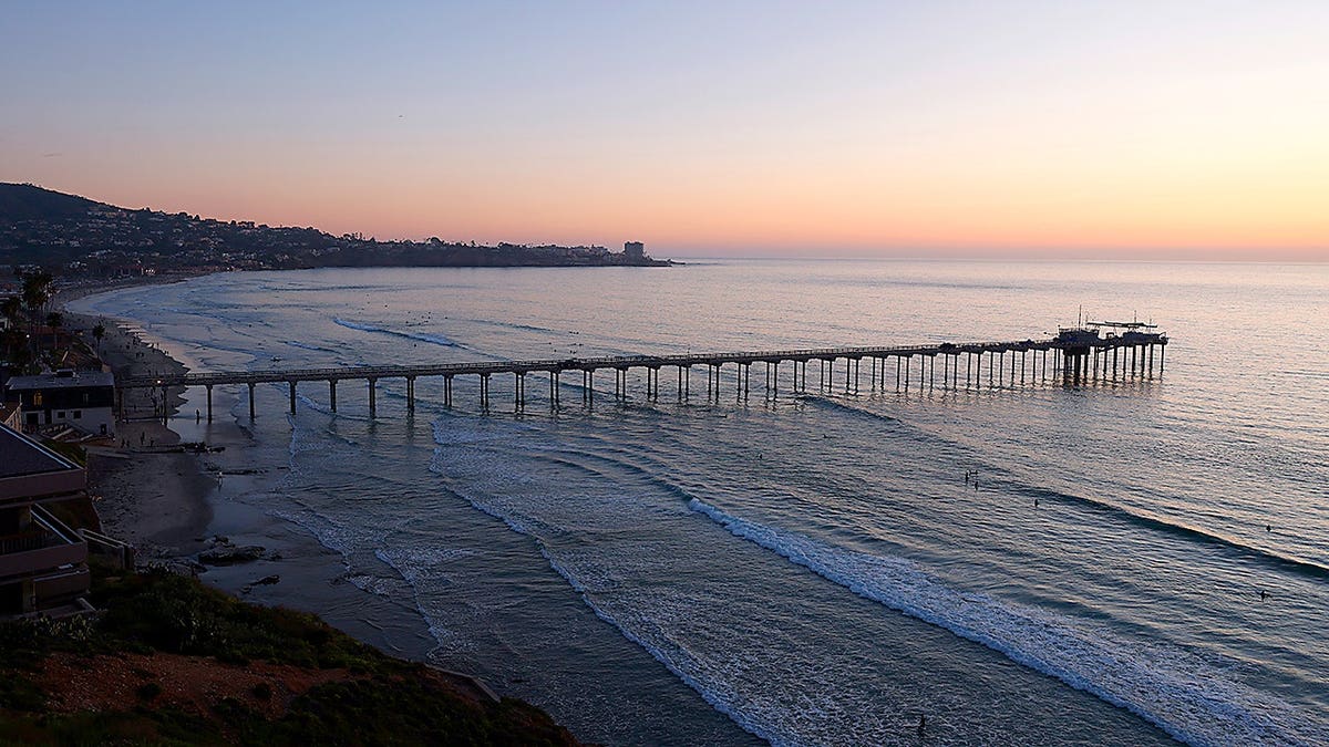 A wide view of the Ellen Browning Scripps Memorial Pier extending into the Pacific Ocean at La Jolla Shores Beach in San Diego, California, during sunset.