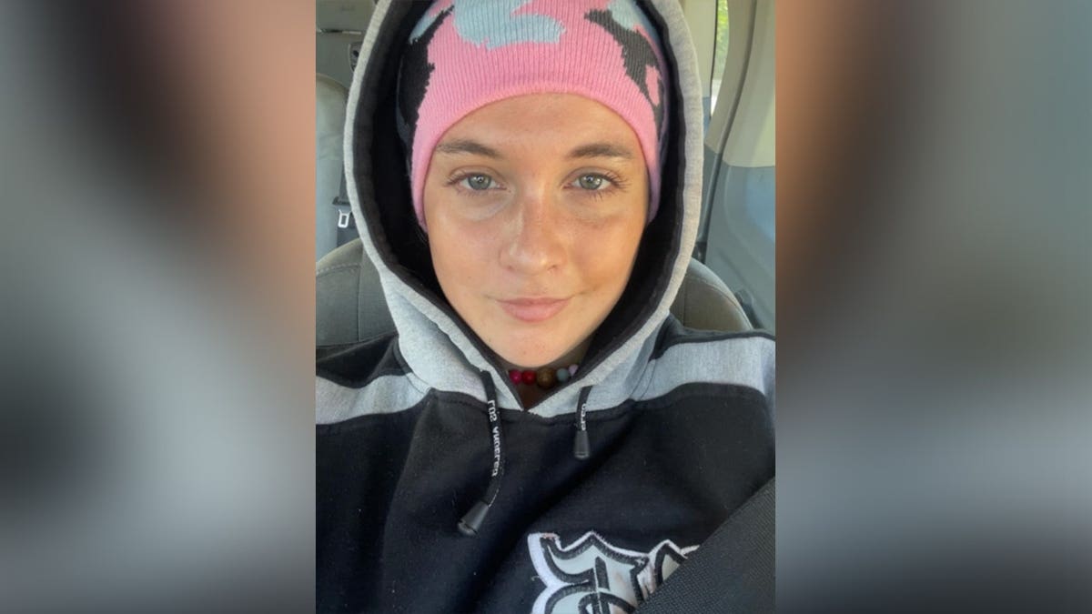 A selfie of Bryn Waite wearing a black-and-gray hoodie and a pink knit beanie while seated inside a vehicle.