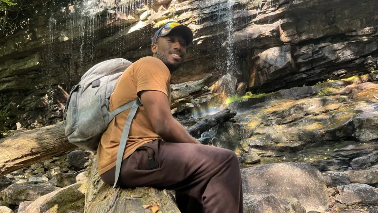 A man sitting on a log in front of a rocky waterfall, wearing a brown t-shirt and gray backpack.