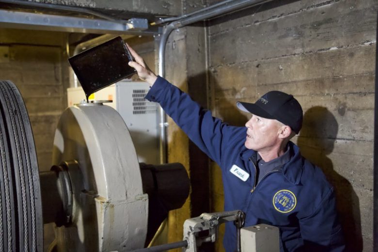 A technician in a blue jacket and cap inspects machinery in an industrial setting, holding a tablet device above a large metal component.