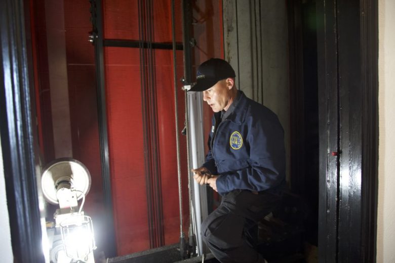 A man in uniform inspects elevator machinery near a lit work lamp inside an elevator shaft.