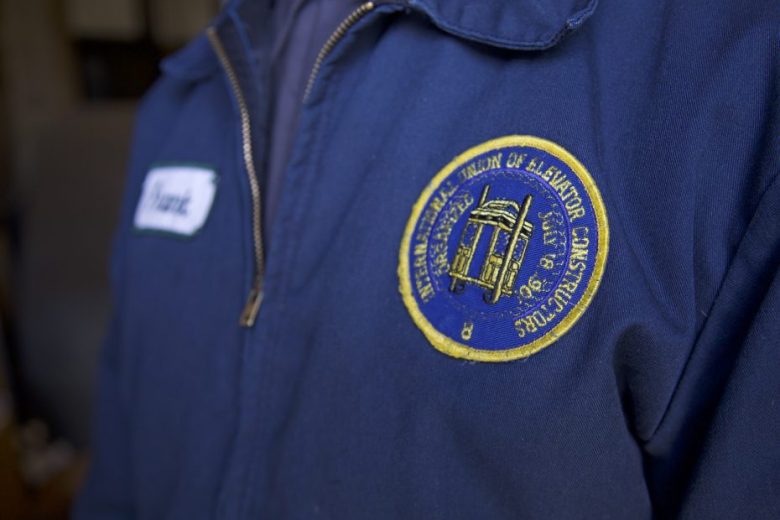 Close-up of a navy blue jacket with a circular patch reading "International Union of Elevator Constructors," worn by a person with a partially visible name tag.