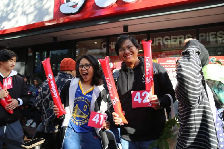 People holding numbered cards and red Jollibee sticks stand and smile outside a Jollibee restaurant during an event.