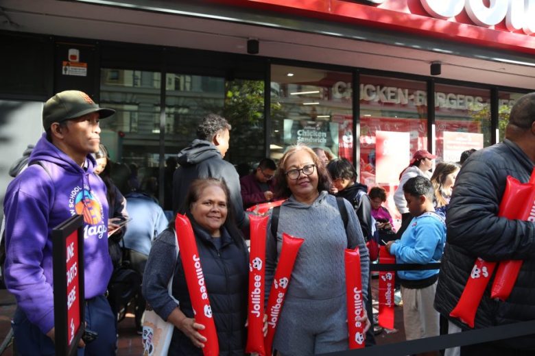 A group of people stand in line outside a Jollibee restaurant, some holding red inflatable sticks with the Jollibee logo.