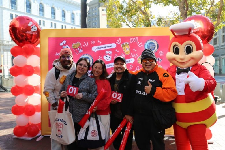 Five people pose with bags and numbered cards in front of a Jollibee backdrop and mascot outdoors; red and white balloons frame the scene.