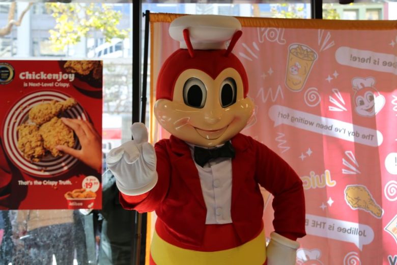 A person in a Jollibee mascot costume stands indoors next to a promotional poster and a decorative backdrop, smiling and posing with one hand raised.