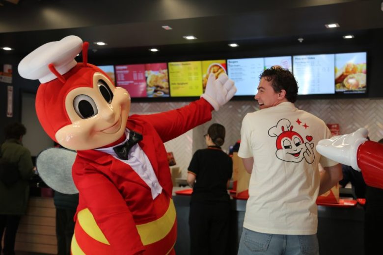 A person in a Jollibee mascot costume poses with a smiling man wearing a Jollibee-themed shirt inside a fast food restaurant.
