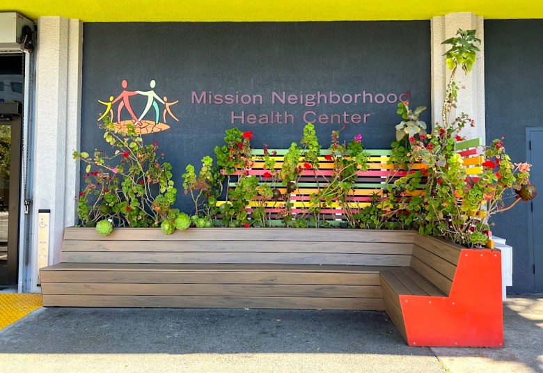 A bench and flowers in front of a wall with the sign "Mission Neighborhood Health Center."