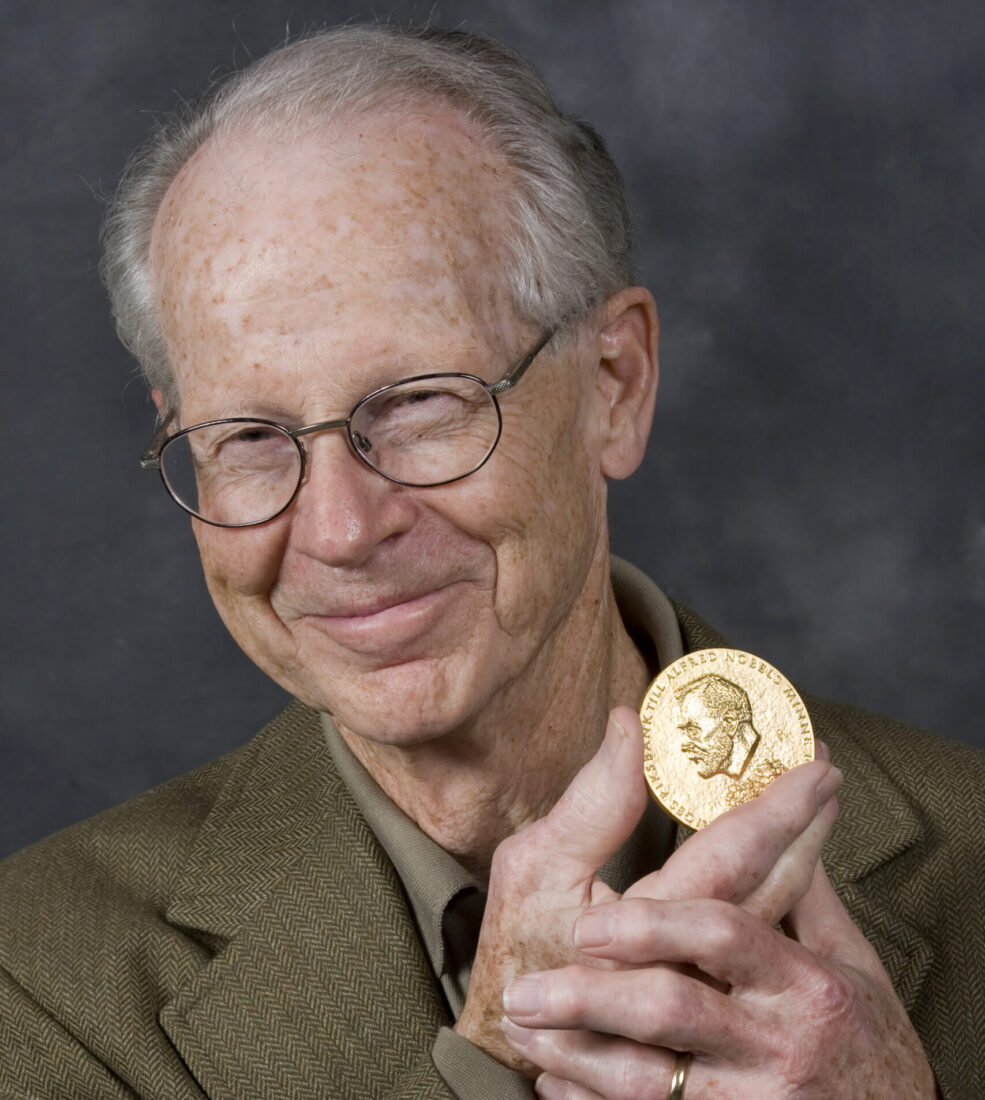 Photo of a smiling bespectacled man holding a gold Nobel coin