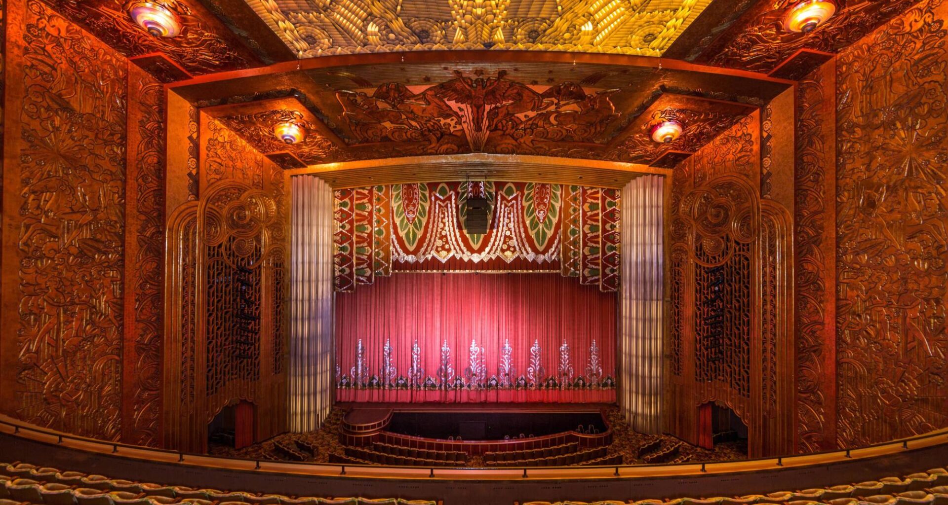 The inside of an ornate movie theater, shown from the balcony, with a large red curtain and seats