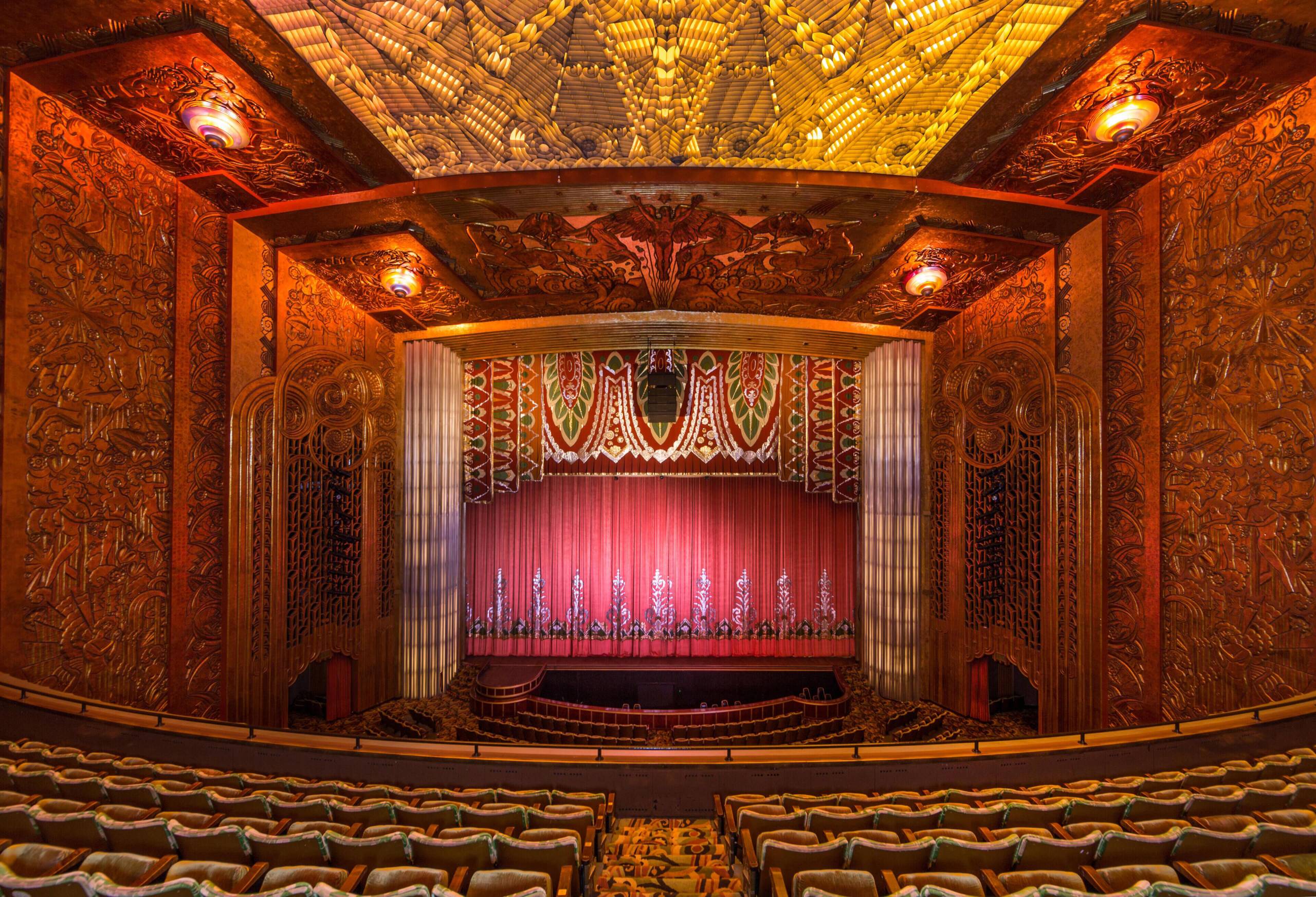 The inside of an ornate movie theater, shown from the balcony, with a large red curtain and seats