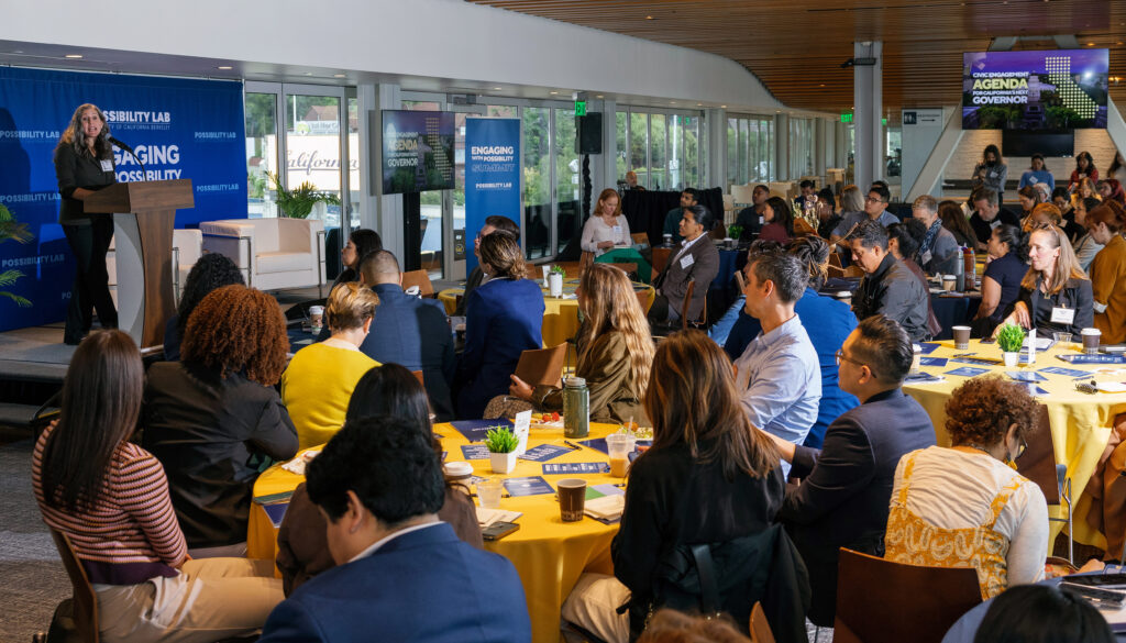 Amy E. Lerman, at a lectern far left, talks to the high-level audience at about 10 tables during the Engaging with Possibility Summit