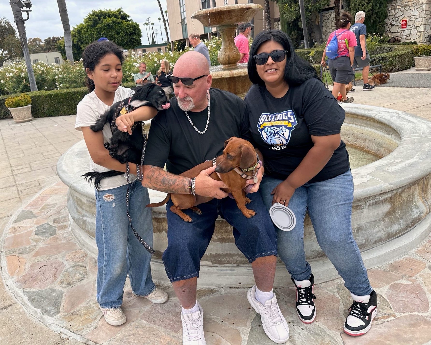 Two adults and a child sit outside on a stone bench while holding small dogs.