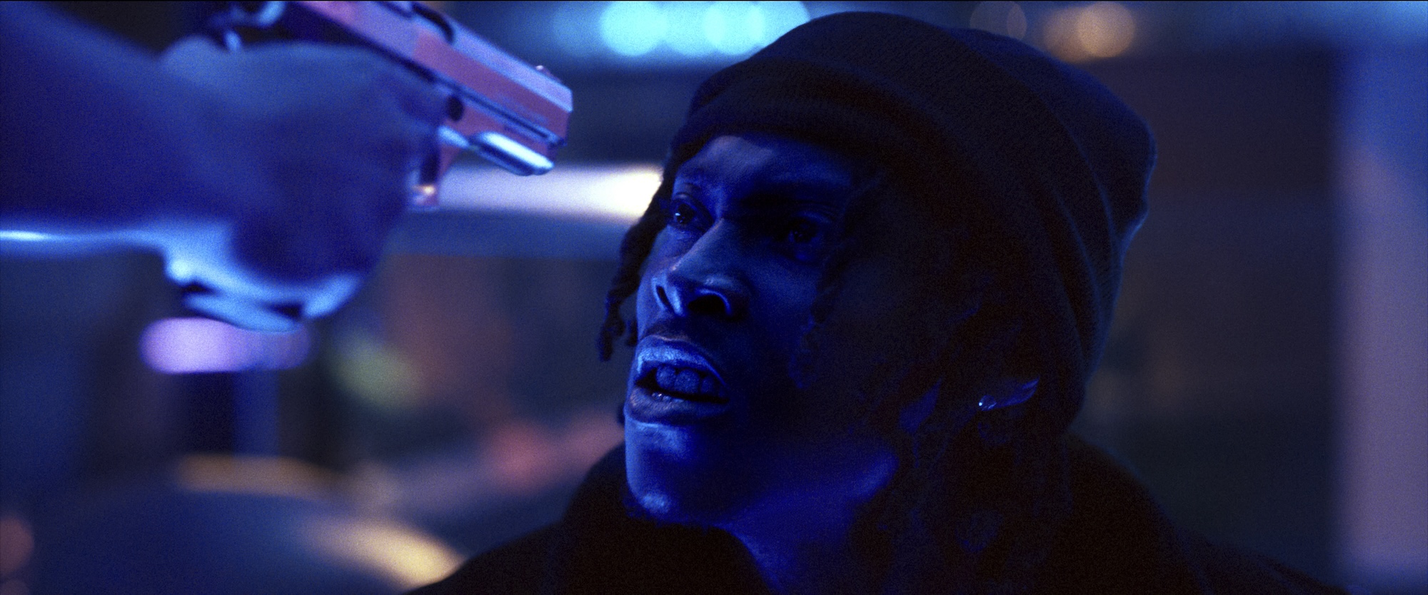 A young Black man in a beanie has a terrified look as a pistol is aimed at his face in a darkly-lit room