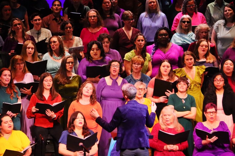 A group of women in an array of brightly-colored shirts sings in a chorus, with many of them holding songbooks. In front of them, with her back to the camera, is a woman wearing a purple blazer.