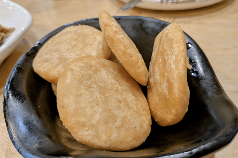 A black Cantoo dish holds five round, golden-brown fried bread pieces on a beige table surface.