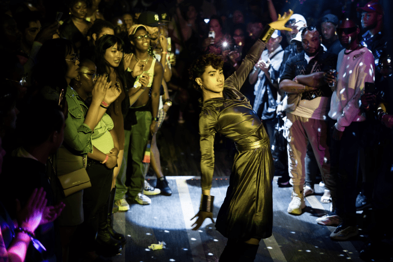 A performer in a dark, shiny outfit strikes a dramatic pose in a dimly lit room, surrounded by a lively, cheering crowd.