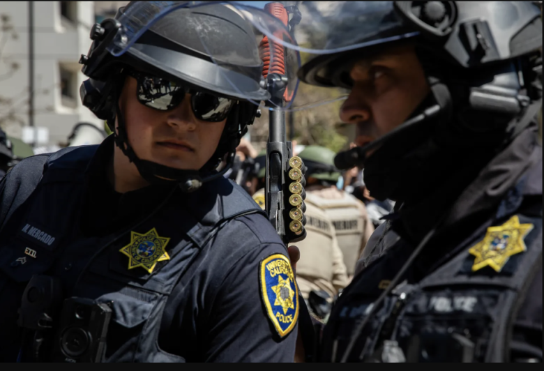 Two police officers in helmets and sunglasses stand closely. One holds a baton with cartridges. Badges read "University of California Police."