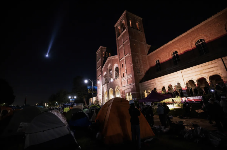 Tents line a campus lawn at night with a large brick building illuminated in the background. A searchlight beams across a dark sky.