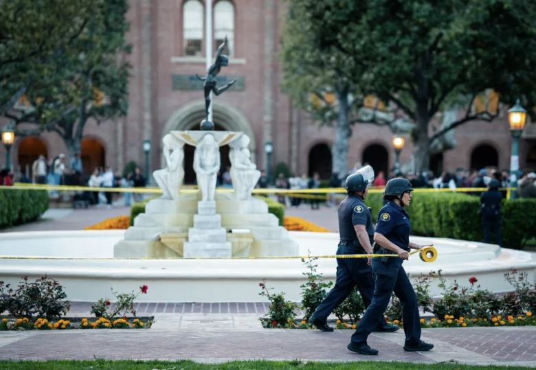 Police officers place yellow caution tape around a decorative fountain with sculptures, in front of a large brick building. A crowd watches nearby.
