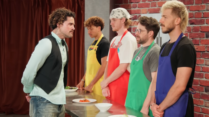 five men on a stage: one is in a vest, the others are in aprons of different colors