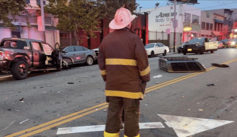 A firefighter in uniform stands in the street near two damaged vehicles and debris after a collision, with emergency lights and responders visible in the background.