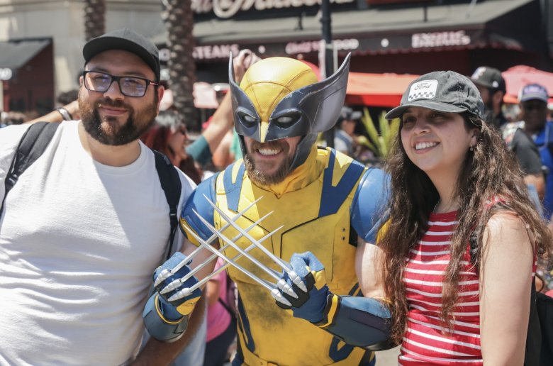 A man in the yellow-and-blue costume of the X-Man Wolverine (center) holds up his claws while posing with two people in regular clothing.