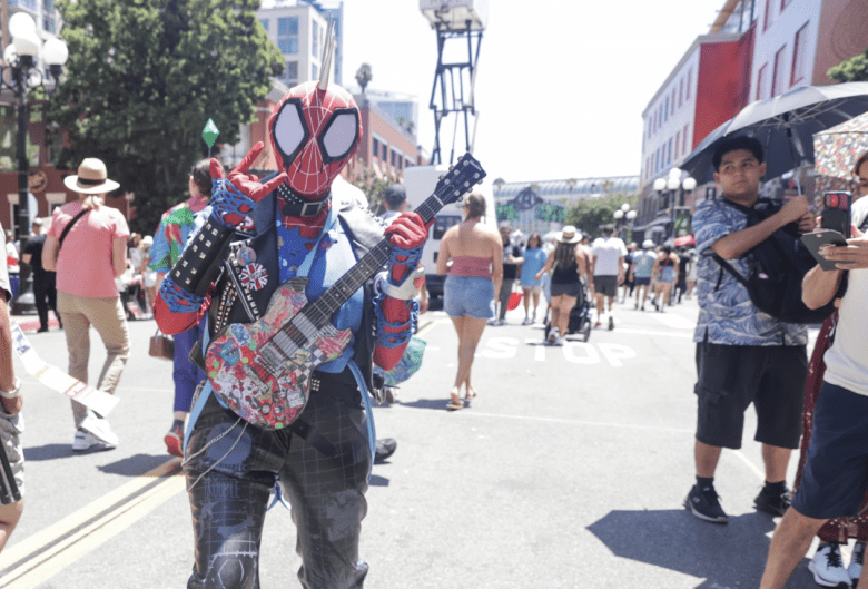 A cosplayer wearing a distinctive Spider-Man mask with a mohawk throws up the "rocker horns" finger pose. They are wearing a leather vest on their costume and carrying a small electric guitar.
