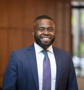 A headshot photo of Shaun Ossei-Owusu smiling while wearing a dark blue suit jacket and purple tie.