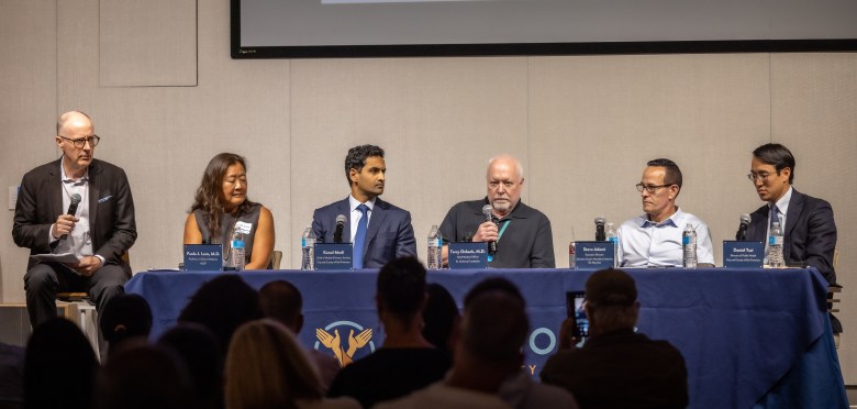 Six people sit on stage before an audience at a public forum.