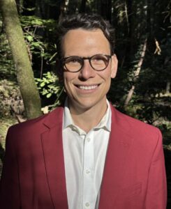 A headshot photo of Steven Bazley wearing eyeglasses and a dark rasberry-colored suit jacket.