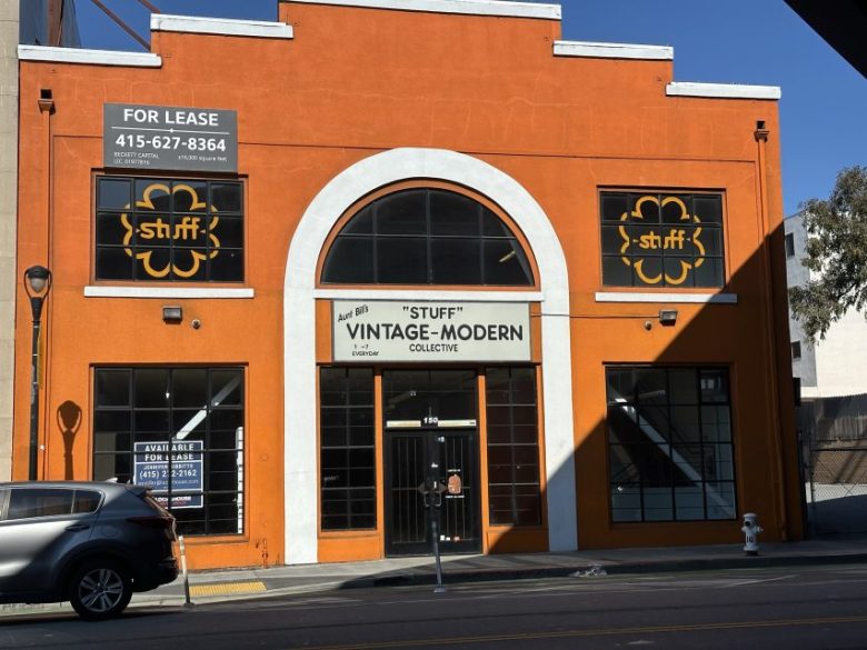 Orange-painted building with large arched window, signs for "Stuff Vintage-Modern Collective," and "For Lease" banners, viewed from the street with a parked car in front.