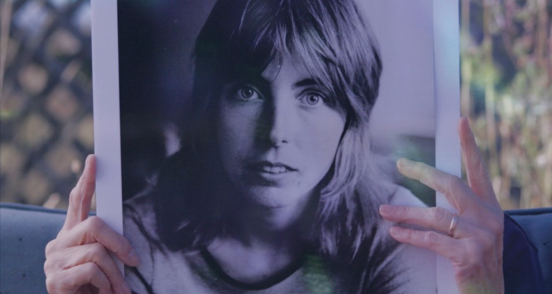 A senior person on a couch holds up a large, black and white photo of a young woman's face. The image hides the face of the person on the couch.