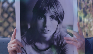 A senior person on a couch holds up a large, black and white photo of a young woman's face. The image hides the face of the person on the couch.