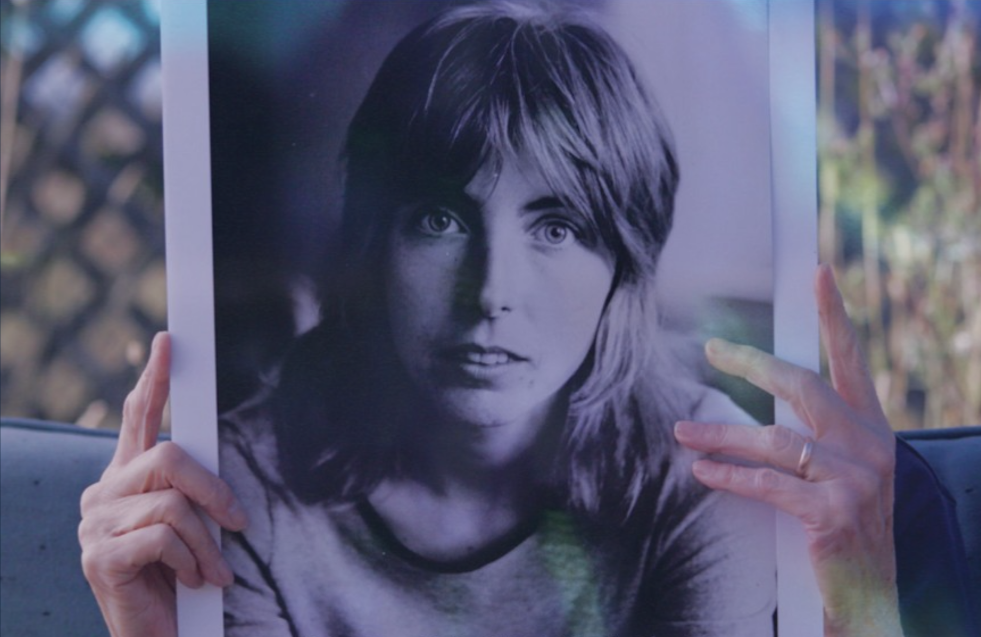 A senior person on a couch holds up a large, black and white photo of a young woman's face. The image hides the face of the person on the couch.