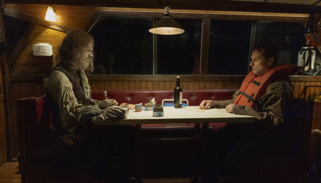Widow’s Bay Mayor Loftis sits opposite the town's tattered, 300-year-old unburied founder inside a dimly lit boat, capturing the show's strange mix of supernatural dread and deadpan humor.