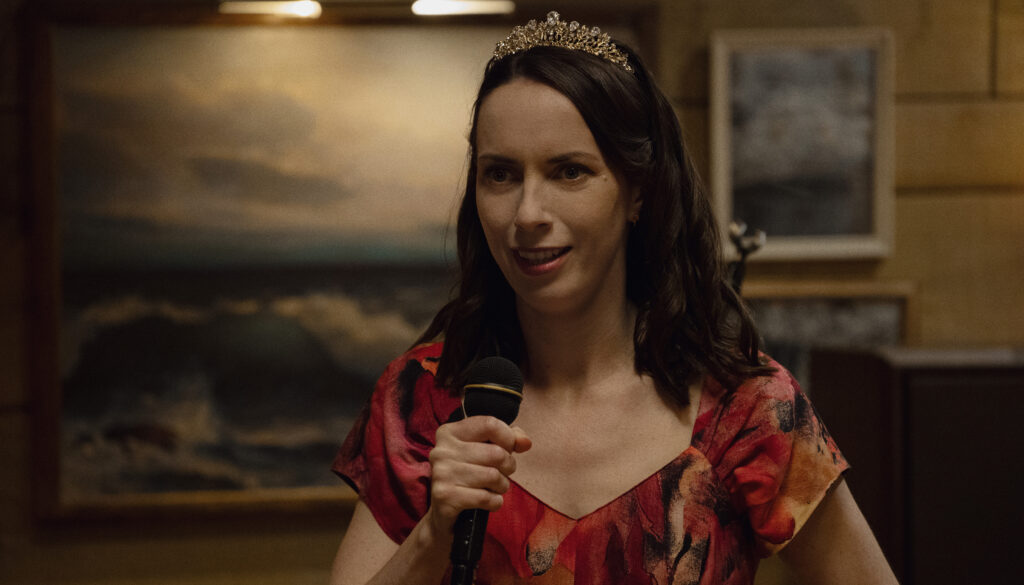 Widow’s Bay character Patricia wearing a tiara and holding a microphone, radiating a relatable mixture of awkward anxiety and determination.
