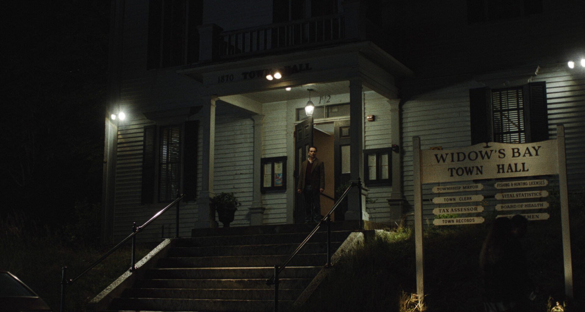 Still from Widow’s Bay showing Mayor Tom Loftis framed in the illuminated doorway of a dark, eerie town hall, beside a wooden sign listing local municipal services.