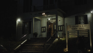 Still from Widow’s Bay showing Mayor Tom Loftis framed in the illuminated doorway of a dark, eerie town hall, beside a wooden sign listing local municipal services.