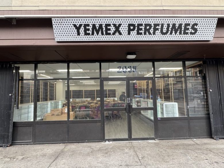 Storefront with large glass windows and a sign reading "YEMEX PERFUMES" above the entrance. Shelves and boxes are visible inside the brightly lit shop.