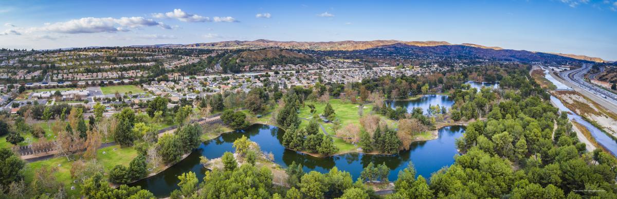 Yorba Regional Park in Anaheim featuring a wide aerial view of scenic lakes, tree-lined green spaces, walking paths, and nearby neighborhoods with rolling hills in the distance under a clear blue sky.
