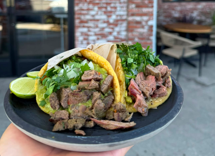 Two tacos de bistec from Taqueria Amor a Mi, on a plate being held up by a hand, two wedges of lime on the side