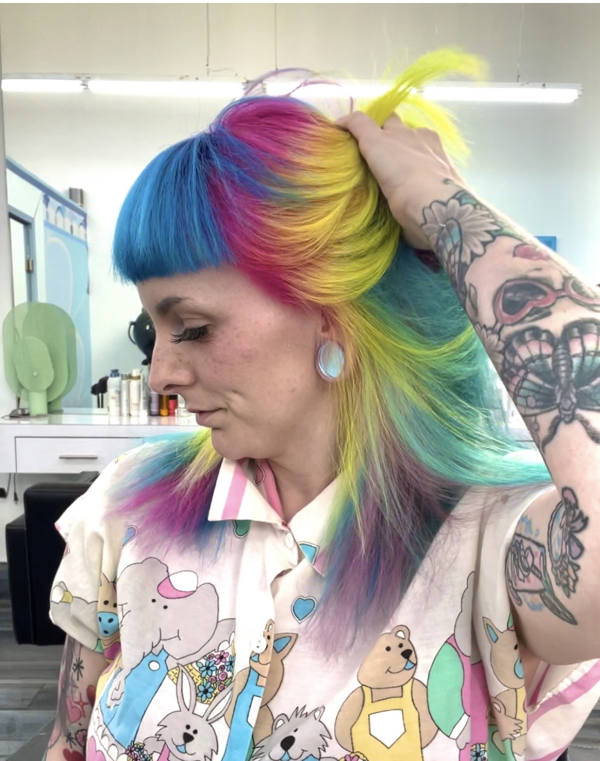 Woman with rainbow-colored hair, tattoos, and earrings, looking down in a room with shelves and objects in the background.