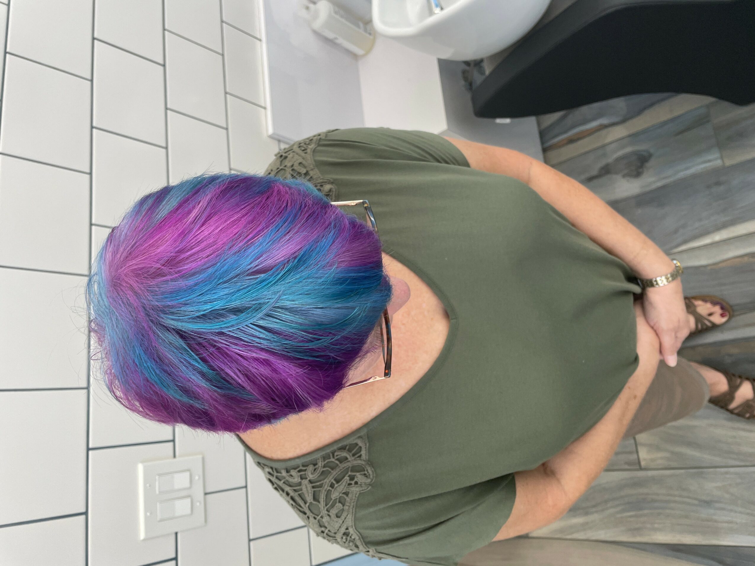 Person with colorful hair in shades of pink, purple, and blue, wearing a green top, standing in a tiled bathroom.