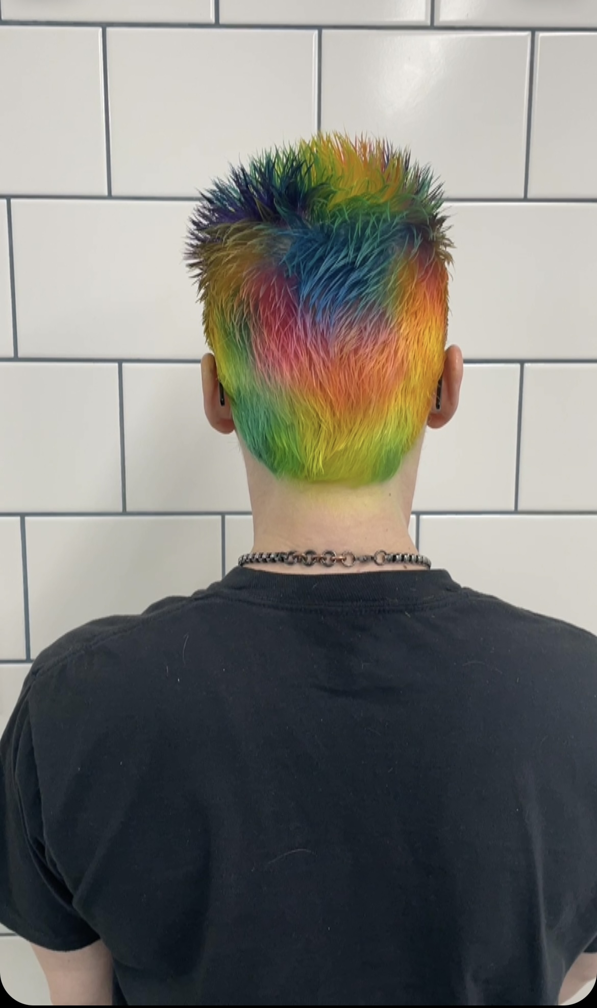 Person with rainbow-colored spiked hair, facing away, in front of white tiled wall, wearing a black shirt and necklace.