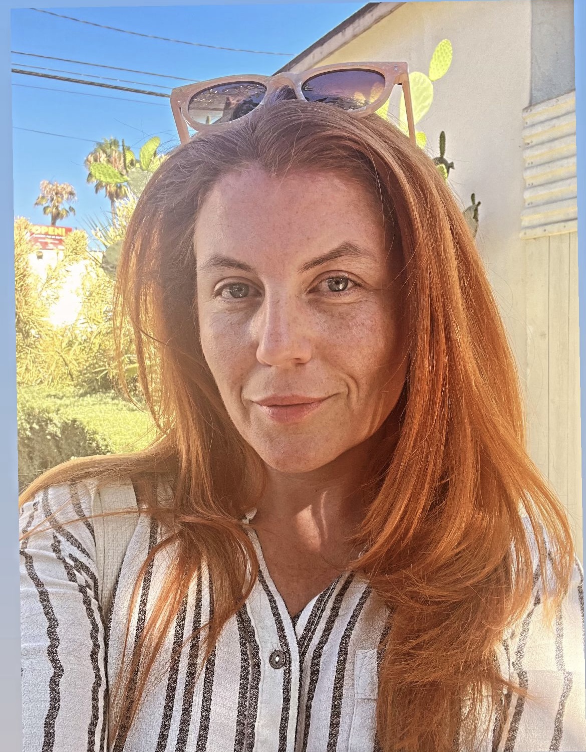 Woman with long red hair outdoors, wearing sunglasses on head and striped shirt, smiling at camera.